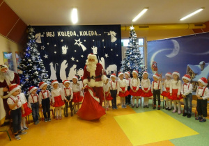 Mikołaj z workiem prezentów stoi pomiędzy dziećmi, w tle świąteczna dekoracja.